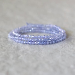 May include: A multi-strand bracelet composed of small, faceted, light periwinkle blue beads. The beads are arranged in a circular, coiled formation, reflecting light across their numerous cut surfaces to create a shimmering, textured appearance.