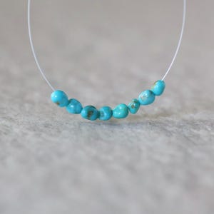 May include: A delicate necklace featuring seven small, polished turquoise beads strung on a thin, clear nylon cord. The beads are irregular in shape, displaying vibrant shades of blue with subtle brown matrix inclusions, arranged in a gentle curve.