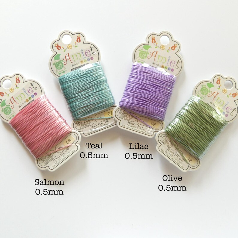 May include: Four spools of Amiet thread in salmon, teal, lilac, and olive colors. Each spool is labeled with the color name and 0.5mm thickness. The packaging features a floral design and the Amiet logo.