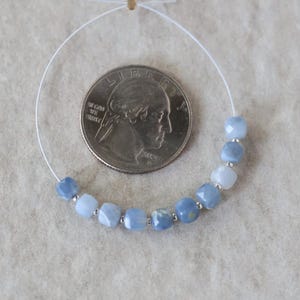 May include: A strand of ten faceted, light blue square beads separated by small silver-toned spacers, arranged in a curve next to a United States quarter coin for scale. The beads exhibit varying shades of blue and a semi-translucent appearance.