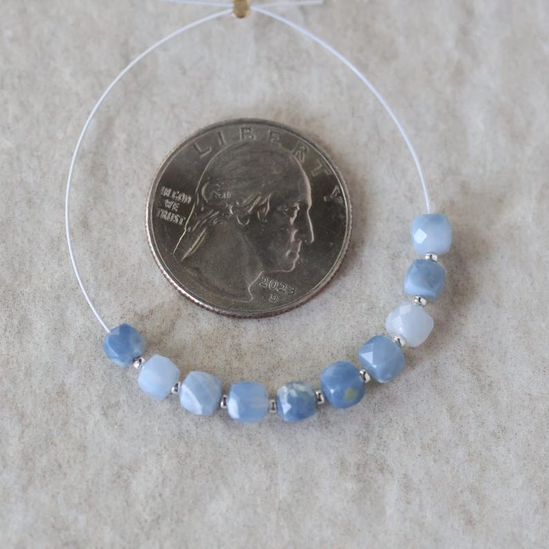 May include: A strand of ten faceted, light blue square beads separated by small silver-toned spacers, arranged in a curve next to a United States quarter coin for scale. The beads exhibit varying shades of blue and a semi-translucent appearance.