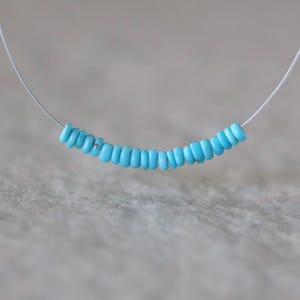 May include: A delicate necklace featuring a single row of small, round, bright turquoise beads strung on a thin, clear, flexible cord. The beads are arranged in a gentle curve, creating a simple and clean design that rests along the neckline.