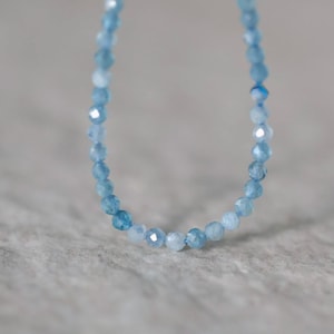 Puede incluir: Un delicado collar compuesto por peque&ntilde;as cuentas de piedras preciosas facetadas en color azul claro. Las cuentas var&iacute;an ligeramente de tono, desde un azul cielo p&aacute;lido hasta un azul cer&uacute;leo m&aacute;s profundo, dispuestas en una l&iacute;nea curva continua que resalta sus superficies reflectantes.