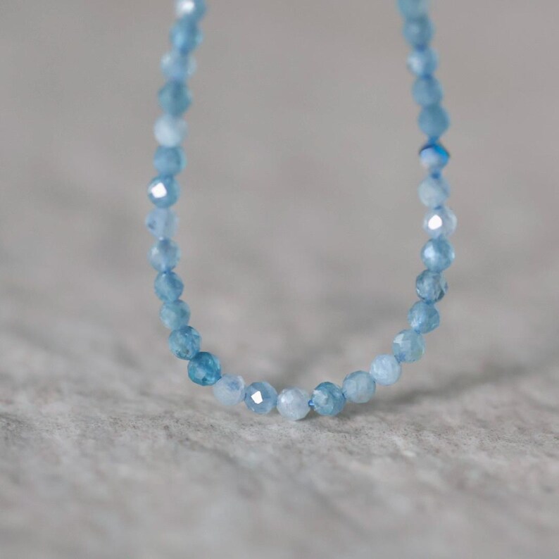 Puede incluir: Un delicado collar compuesto por peque&ntilde;as cuentas de piedras preciosas facetadas en color azul claro. Las cuentas var&iacute;an ligeramente de tono, desde un azul cielo p&aacute;lido hasta un azul cer&uacute;leo m&aacute;s profundo, dispuestas en una l&iacute;nea curva continua que resalta sus superficies reflectantes.