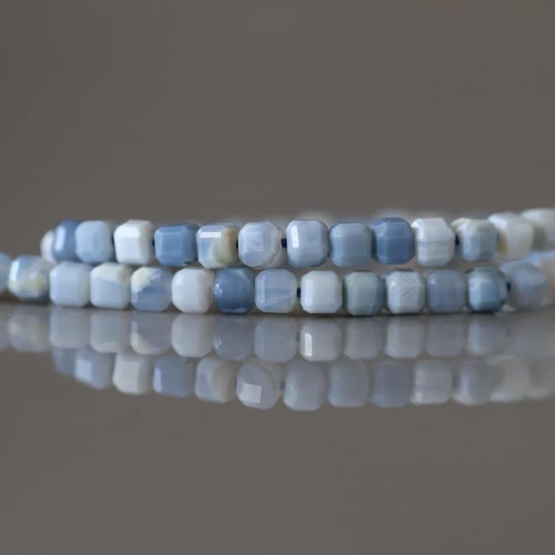 Puede incluir: Primer plano de dos hilos de cuentas de piedras preciosas facetadas de color azul claro y blanco. Las cuentas cuadradas reflejan la luz, creando un efecto brillante. Las cuentas est&aacute;n ensartadas, lo que sugiere que son para la fabricaci&oacute;n de joyas o manualidades.