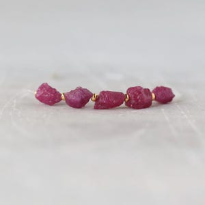 May include: A bracelet featuring five raw, deep pink ruby gemstones arranged in a horizontal line. The stones are separated by small, polished gold-toned metal beads, creating a delicate contrast between the rough texture and the metallic finish.