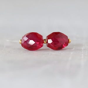 May include: Two faceted, deep red, oval-shaped beads are strung together on a thin, clear filament. A small, polished gold-toned metal spacer bead is positioned between the two red beads, creating a simple, symmetrical jewelry component.
