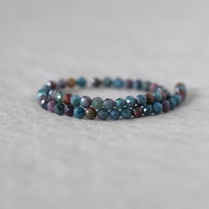 May include: A strand of small, faceted, round beads in shades of teal, blue, and deep reddish-brown. The beads are arranged in a circular, coiled pattern, showcasing a reflective, multi-faceted surface texture across each individual bead.