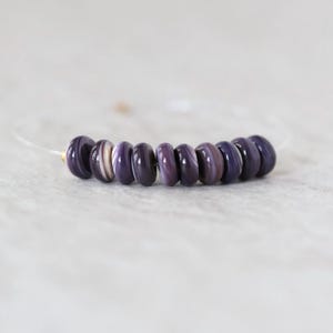 May include: A bracelet featuring ten smooth, round, disc-shaped beads in varying shades of deep purple and lavender with subtle white swirling patterns. The beads are strung on a thin, clear, flexible cord that maintains a circular shape.