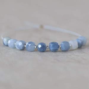 May include: A bracelet featuring a series of faceted, light blue gemstone beads interspersed with small, polished silver-tone metal spacer beads. The beads are strung on a thin, white cord that forms a circular loop, showcasing a cool-toned palette.