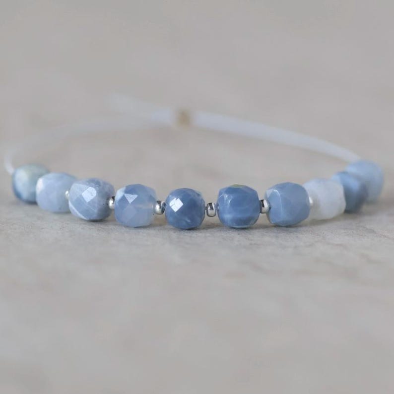 May include: A bracelet featuring a series of faceted, light blue gemstone beads interspersed with small, polished silver-tone metal spacer beads. The beads are strung on a thin, white cord that forms a circular loop, showcasing a cool-toned palette.