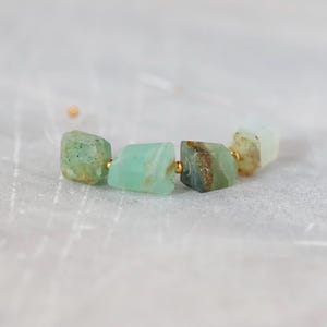 May include: A bracelet featuring four raw, irregularly shaped green stones with brown inclusions, separated by small gold-toned metal beads. The stones have a semi-translucent appearance and a rough, natural texture, strung together on a thin cord.
