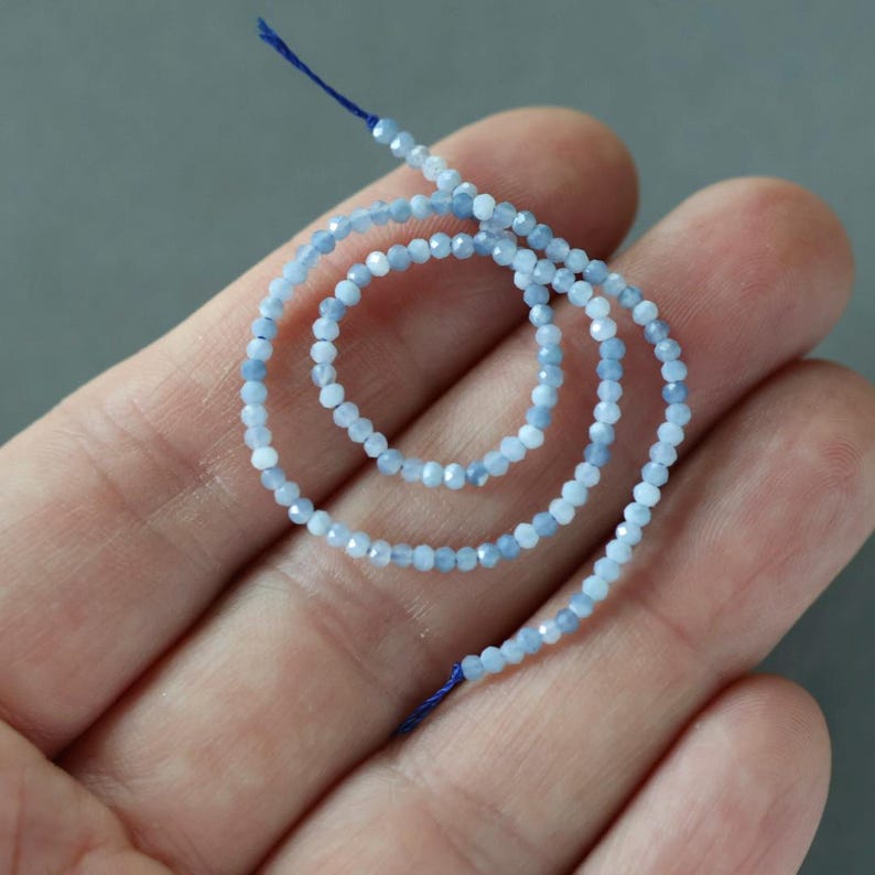 May include: A strand of light blue faceted beads, strung on a dark blue thread. The beads are small and have a subtle sparkle. The strand is held in a hand against a gray background. Ideal for jewelry making.