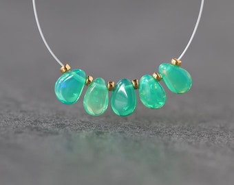 Könnte beinhalten: Eine Halskette mit fünf glatten, tropfenförmigen grünen Opalperlen, die in einer Reihe angeordnet sind. Jeder Opal ist durch eine kleine, polierte goldfarbene Metallzwischenperle getrennt. Die Perlen sind auf einem dünnen, klaren Monofilamentfaden.