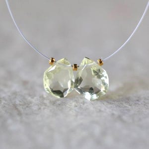 May include: Two faceted, pale yellow, pear-shaped gemstones are strung on a thin, clear filament. The stones are separated and flanked by small, metallic gold-colored spacer beads, creating a delicate, symmetrical arrangement for jewelry.
