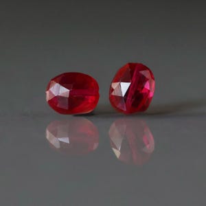 May include: Two faceted, oval-shaped, deep red gemstones. The gems have a high luster and reflect light, creating bright highlights. The background is a dark gray, providing contrast to the vibrant red stones. The gems are likely for jewelry making.