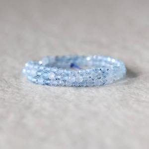 May include: A delicate ring composed of small, faceted light blue beads arranged in a circular band. The beads have a translucent quality and a reflective surface, creating a subtle sparkle. A thin blue thread is visible within the bead structure.