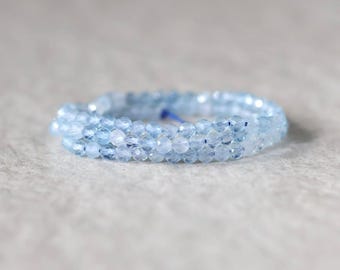 May include: A delicate ring composed of small, faceted light blue beads arranged in a circular band. The beads have a translucent quality and a reflective surface, creating a subtle sparkle. A thin blue thread is visible within the bead structure.