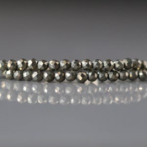 May include: Two strands of faceted pyrite beads. The beads are a metallic gray color and have a reflective surface. The beads are strung together, creating a necklace or bracelet. The background is a soft gray.