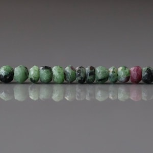 May include: A strand of natural gemstone beads. The beads are a mix of green, black, and pink colors. The beads are roughly cylindrical in shape and have a slightly rough texture. The beads are strung together on a thin, dark string.
