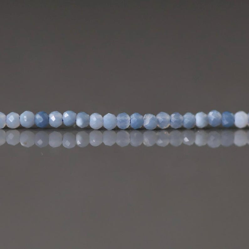 May include: A strand of faceted, light blue gemstone beads. The beads are small, round, and evenly spaced, reflecting light. The background is a gradient of gray.