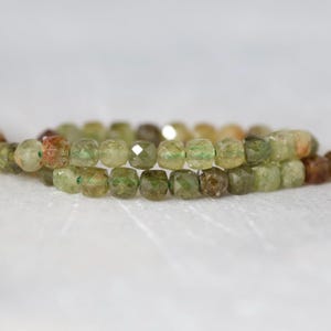 May include: A strand of faceted, translucent beads in varying shades of olive green, moss green, and brownish-gold. The small, rounded beads are strung together, displaying a reflective, multi-faceted surface that catches light across the entire length.