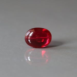 May include: A translucent, oval-shaped, deep red gemstone. The polished surface reflects light, creating highlights and shadows. The gem sits on a reflective surface, mirroring its form.