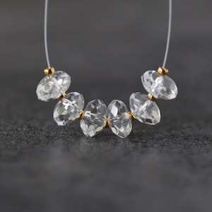 May include: A strand of six clear, faceted crystal beads arranged in a curved line on a thin, transparent filament. Each crystal bead is separated by a small, round, metallic gold-toned spacer bead, creating a repeating pattern of clear and gold.