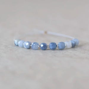 May include: A bracelet featuring a series of faceted, light blue gemstone beads interspersed with small, round silver-toned metal spacers. The beads are strung on a thin, white cord that extends from both sides of the central beaded section.