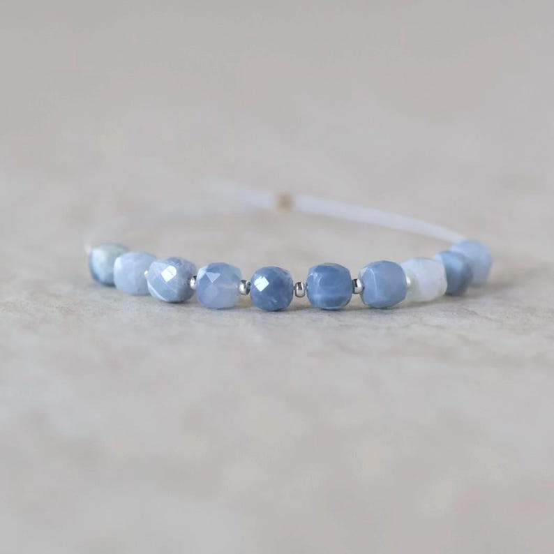 May include: A bracelet featuring a series of faceted, light blue gemstone beads interspersed with small, round silver-toned metal spacers. The beads are strung on a thin, white cord that extends from both sides of the central beaded section.