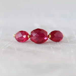 May include: Three faceted, deep red beads arranged in a horizontal row. The central bead is larger than the two flanking beads. Small, polished gold-toned metal spacers are positioned between each of the three translucent red beads.
