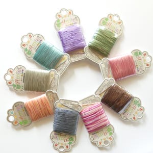 May include: A collection of colorful embroidery floss spools arranged in a circular pattern. The spools are in various shades, including teal, lavender, green, pink, and brown. Each spool is labeled with the brand name "Amiet".