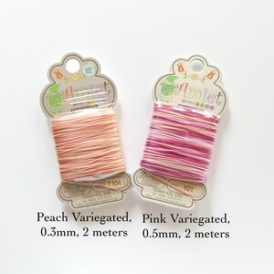 May include: Two spools of Amiet thread are displayed. One is peach variegated, 0.3mm thick, and the other is pink variegated, 0.5mm thick. Each spool contains 2 meters of thread. The packaging includes Japanese text and the brand name.