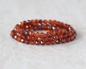 Könnte beinhalten: Ein kreisförmiges Armband aus mehreren Strängen kleiner, facettierter, tief rotbrauner Granatperlen. Die Perlen sind in einer durchgehenden Schlaufe angeordnet und reflektieren das Licht auf ihren vielen geschliffenen Oberflächen.