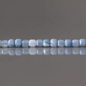 Puede incluir: Un collar de cuentas cuadradas de color azul claro. Las cuentas tienen una calidad ligeramente transl&uacute;cida, con sutiles variaciones de color y textura. Las cuentas est&aacute;n ensartadas, creando una l&iacute;nea continua. El fondo es gris neutro.