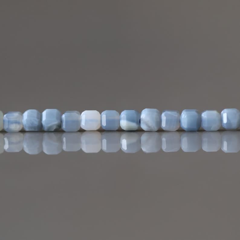 Puede incluir: Un collar de cuentas cuadradas de color azul claro. Las cuentas tienen una calidad ligeramente transl&uacute;cida, con sutiles variaciones de color y textura. Las cuentas est&aacute;n ensartadas, creando una l&iacute;nea continua. El fondo es gris neutro.