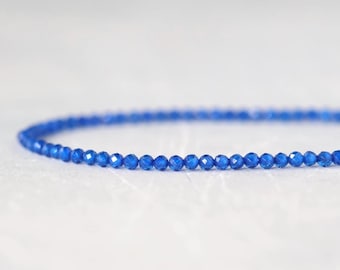Könnte beinhalten: Eine einzelne Schnur aus kleinen, facettierten, tiefblauen Perlen, die in einer sanften Kurve angeordnet sind. Jede einzelne Perle hat eine reflektierende, mehrfach facettierte Oberfläche, die das Licht einfängt und eine lebendige Farbe erzeugt.