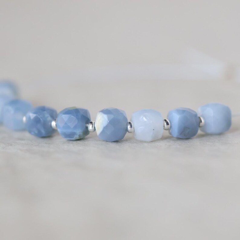 May include: A strand of faceted, light blue gemstone beads arranged in a row. Each bead is separated by a small, round, metallic silver spacer bead. The beads exhibit varying shades of pale blue and white, with a polished, multi-faceted surface.