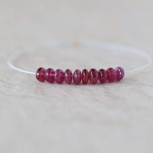 May include: A delicate bracelet featuring a single row of ten small, round, deep pinkish-red gemstone beads strung on a thin, white cord. The beads are uniform in size and shape, displaying a smooth, polished surface with subtle variations in color.