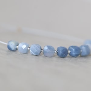 May include: A bracelet featuring a series of faceted, light blue, cube-shaped beads strung on a thin white cord. Small, round, metallic silver spacer beads are positioned between each of the blue stone beads, creating a repeating pattern.