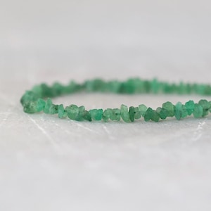 May include: A bracelet composed of small, irregular, raw green stone chips strung together in a circular shape. The stones exhibit varying shades of vibrant green and have a textured, unpolished appearance, creating a continuous band of natural mineral fragments.