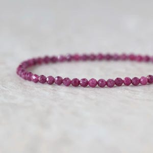 May include: A delicate bracelet composed of small, round, faceted magenta-colored beads. The beads are uniform in size and arranged in a single continuous strand, showcasing a reflective, textured surface that catches the light.