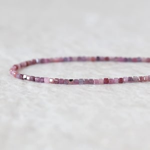 May include: A delicate strand of small, faceted cube-shaped beads in varying shades of pink, ranging from light blush to deep magenta. The beads are strung together in a single, continuous line, showcasing a subtle gradient of pink tones.