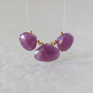 May include: Three faceted, teardrop-shaped purple gemstones strung on a thin, clear filament. The stones are separated by small, round, metallic gold-colored beads, creating a delicate arrangement of purple and gold jewelry components.