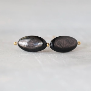 May include: Two oval-shaped black beads with a subtle metallic sheen and visible internal striations, connected by a small gold-toned spacer bead and featuring gold-toned metal loops at the outer ends of the bead arrangement.