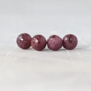 May include: Four round, faceted beads in a deep, dark magenta-red color. Each bead features a textured, multi-faceted surface that reflects light, arranged in a horizontal line on a light, neutral-toned surface.