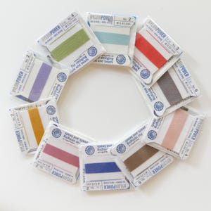 May include: A collection of nylon stringing cords in various colors, including green, blue, red, and pink. Each cord is packaged in a clear plastic sleeve with the "NYLONPOWER" logo and specifications for stringing pearls and beads. The cords are arranged in a circular pattern.