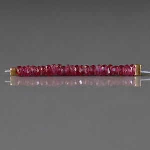 May include: A strand of faceted, deep red ruby beads. The beads are strung together, with gold-colored end caps. The beads are reflecting light, and the background is a neutral gray.