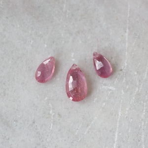 May include: Three pear-shaped pink gemstones with faceted surfaces. The stones are arranged in a loose cluster, displaying a translucent quality and a vibrant rose-pink hue. Each gemstone features a pointed top and a rounded base.
