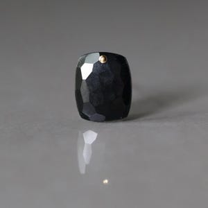 May include: A faceted, black gemstone pendant with a small gold-colored accent. The pendant has a rectangular shape with rounded edges and a polished finish. The gemstone reflects light, creating highlights and shadows.